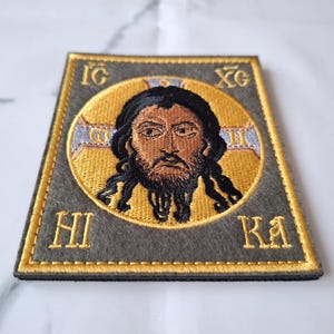 Op de afbeelding: Geborduurde patch met een religieus icoon met een gouden halo en donker haar. De afbeelding staat tegen een grijze achtergrond met gouden letters en randen. De patch heeft de Griekse letters IG, XC, HI en KA.