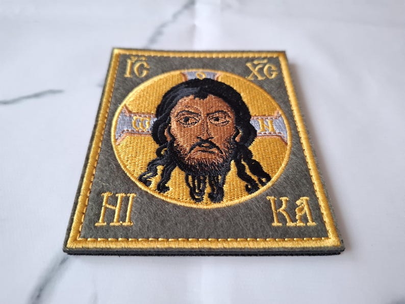 Op de afbeelding: Geborduurde patch met een religieus icoon met een gouden halo en donker haar. De afbeelding staat tegen een grijze achtergrond met gouden letters en randen. De patch heeft de Griekse letters IG, XC, HI en KA.