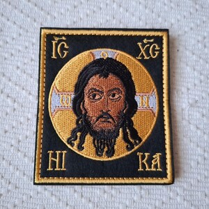 Op de afbeelding: Geborduurde patch met een religieus icoon van een man met donker haar en een baard, tegen een gouden halo. De afbeelding is omlijst in een zwarte vierkant met gouden rand en Griekse letters. Religieus thema.