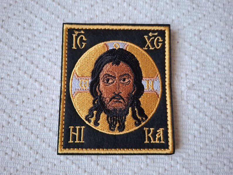 Op de afbeelding: Geborduurde patch met een religieus icoon van een man met donker haar en een baard, tegen een gouden halo. De afbeelding is omlijst in een zwarte vierkant met gouden rand en Griekse letters. Religieus thema.