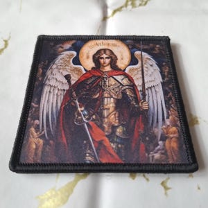 May include: Square patch featuring a detailed illustration of an angel with large white wings, a red cloak, and a sword. The angel has a halo and is set against a dark background. The patch has a black border.