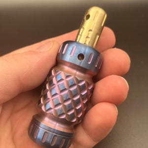 May include: A cylindrical metal tool featuring a textured, diamond-patterned grip section with an iridescent blue and purple finish. The top portion is a smooth, polished brass-colored component with a small circular hole near the rounded tip.