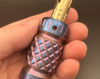May include: A cylindrical metal tool featuring a textured, diamond-patterned grip section with an iridescent blue and purple finish. The top portion is a smooth, polished brass-coloured component with a small circular hole near the rounded tip.