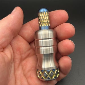 Puede incluir: Un fidget spinner plateado con detalles azules y dorados. Las secciones superior e inferior presentan un patrón de diamantes en azul y dorado. La parte superior tiene un diseño texturizado en oro y azul. El objeto se sostiene en una mano.