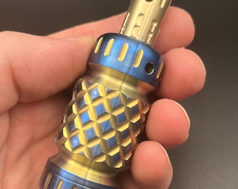May include: A cylindrical metal object featuring a gold-toned top section with a small hole, a central band with a diamond-patterned texture, and blue-toned accents on the top and bottom rings. The surface displays a mix of gold and blue hues.