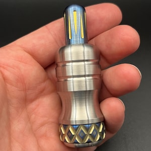 May include: A silver metal object with a blue and gold top and bottom, held in a hand. The top has vertical gold stripes. The bottom has a diamond pattern. The object has a cylindrical shape with a ridged middle section.