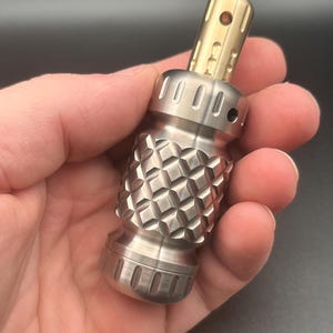 May include: A cylindrical metal tool featuring a brushed silver finish with a textured diamond-patterned grip section. The top portion is a smooth, golden-toned metal cylinder with vertical grooves and a small circular opening near the top edge.