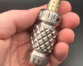 May include: A cylindrical metal object with a brushed silver finish and a textured diamond-patterned centre section. The top features a smooth, gold-toned cylindrical extension with vertical grooves and a small circular opening at the tip.