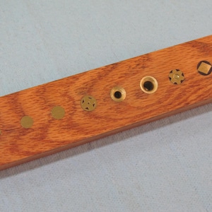 May include: A rectangular wooden block with a reddish-brown finish. It features several small, circular and geometric brass inlays of varying sizes. The inlays are evenly spaced along the length of the block, creating a decorative pattern.