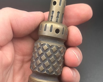 May include: A cylindrical brass lighter featuring a textured diamond-patterned grip in the centre. The top section has a chimney with vertical slots and circular holes, while the base is finished with a series of vertical grooves around the edge.