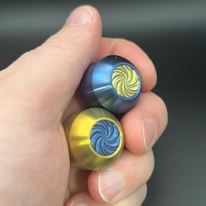 Puede incluir: Dos juguetes fidget de metal se sostienen en una mano. Uno es dorado con un diseño de espiral azul, y el otro es azul con un diseño de espiral dorado. Los juguetes fidget tienen forma redondeada y superficie texturizada.