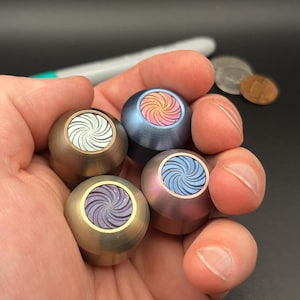 Puede incluir: Una mano sostiene cinco spinners fidget de metal en varios colores: bronce, azul, rosa y morado. Cada spinner presenta un diseño en espiral en un color contrastante. Un bolígrafo y monedas son visibles en el fondo.