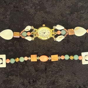 May include: A gold-toned watch with a white face and black numerals. The watch band is adorned with white shell accents, peach, green, and brown beads. The watch band is made of multiple sections.