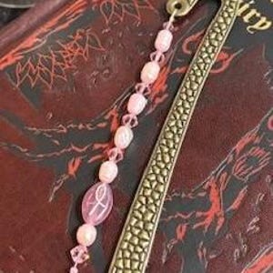 May include: A decorative bookmark with a brass finish and a strand of pink beads. The bookmark has a textured design and a hook at the top. The beaded strand includes oval and round beads.