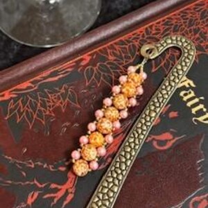 May include: A decorative bookmark with a bronze finish and a beaded strand. The bookmark features a textured, curved design and a strand of orange and pink beads. The bookmark is resting on a book with the word "Fairy" visible.