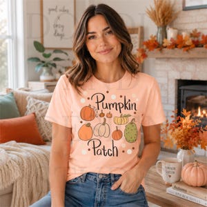 Może przedstawiać: Brzoskwiniowy T-shirt z krótkim rękawem z nadrukiem graficznym przedstawiającym różne dynie w odcieniach pomarańczu, żółci i zieleni. Napis "Pumpkin Patch" wykonano ciemnobrązową czcionką, otoczoną małymi białymi i brązowymi kropkami.
