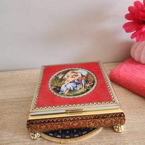 May include: A red and gold metal box with a raised floral design. The lid features a round image of a woman and child in a garden setting.