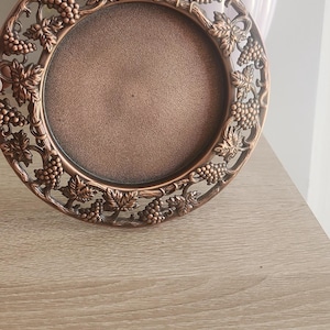 May include: A decorative bronze-colored plate with a grape vine design around the rim. The plate has a circular center and is displayed on a wooden surface. The background includes a vase with pink flowers.