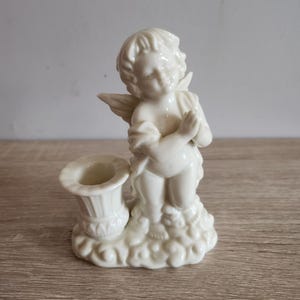 Könnte beinhalten: Ein weißer Kerzenhalter aus Keramik mit einem Putto. Der Putto kniet mit gefalteten Händen neben einem Kerzenhalter. Der Sockel hat ein wolkenartiges Design. Der Kerzenhalter ist ein dekorativer Gegenstand.