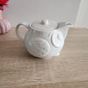 Puede incluir: Tetera de cerámica blanca con forma de búho y tapa. La tetera tiene un diseño de búho detallado con plumas y ojos en relieve.