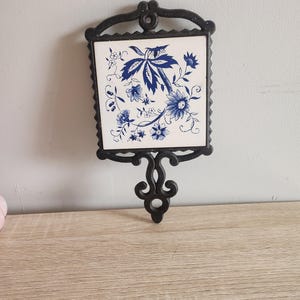 May include: A black cast iron trivet with a white ceramic tile insert featuring a blue floral design.
