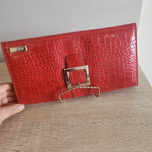 Könnte beinhalten: Eine rote Clutch aus Krokodilleder mit einer goldenen Schnalle und dem Wort "MICHAEL" auf der Vorderseite.