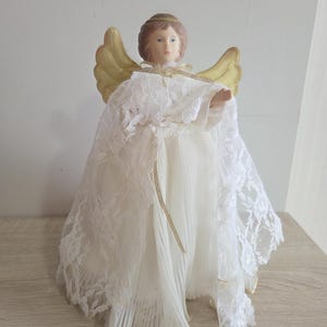 Peut inclure: Figurine d'ange décorative avec une expression sereine. L'ange a des ailes dorées et une robe blanche avec des détails en dentelle. La figurine est posée sur une surface en bois.