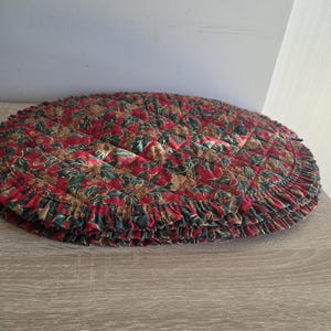 May include: A round, quilted Christmas tree skirt with a ruffled edge. The skirt features a pattern of red, green, and gold floral designs. The skirt is placed on a wooden surface.