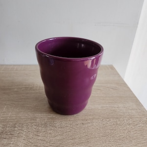 Peut inclure: Une tasse en céramique de couleur violet foncé avec une forme arrondie. La tasse est posée sur une surface en bois brun clair.