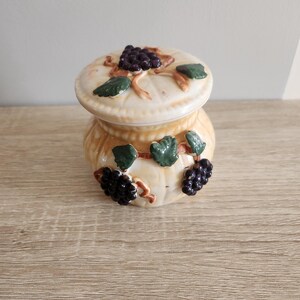 Peut inclure: Un petit pot en céramique avec un couvercle, décoré d'un motif de vigne. Le pot est de couleur crème avec des accents bruns et verts. Le couvercle a un groupe de raisins sur le dessus.