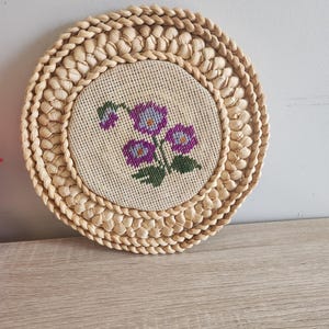 Puede incluir: Un posavasos redondo tejido con un diseño floral en el centro. El posavasos está hecho de materiales naturales y tiene un color marrón claro. El diseño floral presenta flores moradas con hojas verdes.