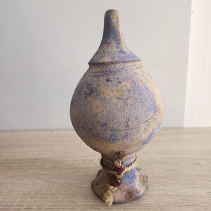 Puede incluir: Un jarrón de cerámica azul y marrón con cuerpo redondeado y cuello estrecho. El jarrón tiene un aspecto rústico y envejecido y está decorado con una cuerda y cuentas alrededor de la base.