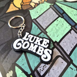 May include: A black and white keychain with the text "LUKE COMBS" in a bold, stylized font. The keychain is attached to a silver metal ring and chain. The background features a colourful, abstract design with green, grey, and blue hues.