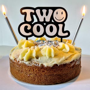 May include: A round cake with white icing and blue and white sprinkles. Two lit candles are in the cake and a black and white cake topper says "TWO COOL" with a smiley face.