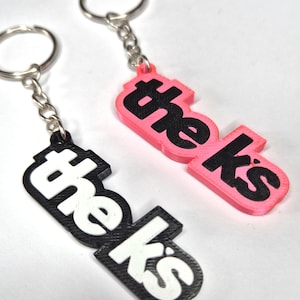 May include: Two keychains with the word "theks" printed on them. One keychain is black with white lettering and the other is pink with black lettering.