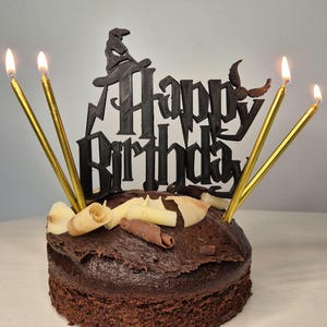 May include: A chocolate cake topped with white and brown chocolate curls. The cake is decorated with a black 3D "Happy Birthday" topper featuring a wizard's hat and a flying broomstick. Five lit gold candles adorn the cake.