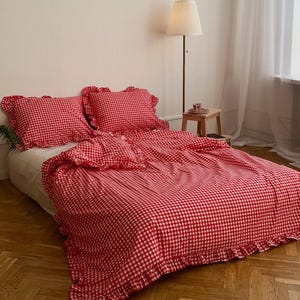 Może przedstawiać: Łóżko z czerwoną i białą poszwą w kratę i pasującymi poszewkami na poduszki. Pościel ma falbanki. Pokój posiada parkiet, roślinę doniczkową, lampę i okno z przezrocznymi zasłonami.