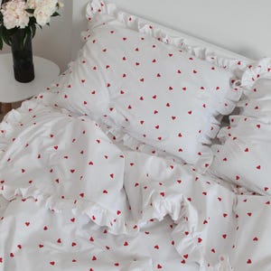 Pode incluir: Conjunto de roupa de cama branco com um padrão de pequenos corações vermelhos. O conjunto inclui uma capa de edredão e fronhas com bordas com babados. Um buquê de flores brancas em um vaso preto está em uma pequena mesa branca.