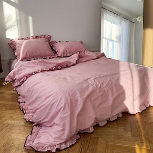 Op de afbeelding: Een roze beddengoedset met gerimpelde randen en donkerrode bies. De set bevat een dekbedovertrek en kussenslopen. Het beddengoed is op een houten vloer gelegd, met een grote boogvormige spiegel op de achtergrond.