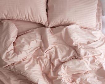 May include: Overhead view of a bed with a light pink striped duvet cover and two matching pillowcases. The bedding is rumpled, creating soft shadows and textures. The overall aesthetic is clean and minimalist, suggesting a focus on comfort and simplicity.