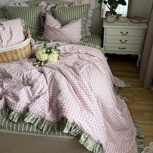 May include: A bed featuring a pink and white gingham duvet cover with ruffled edges, layered over green and white striped sheets. The bedding includes matching gingham and striped pillows, a wicker basket, and a bouquet of white hydrangea flowers.