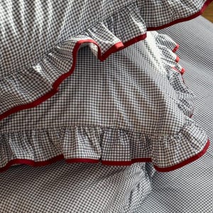 May include: Close-up of a gingham patterned pillowcase with ruffled edges. The fabric is a black and white check pattern with a red trim along the ruffled edges. The pillowcase is part of a bedding set.