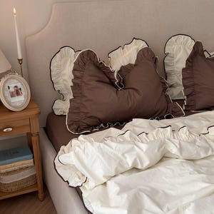 Peut inclure: Un lit avec une literie marron et blanche. Les oreillers sont marron avec des bords à volants blancs et une bordure noire. La housse de couette est blanche avec une bordure noire. Une petite table de chevet en bois avec une photo encadrée et une bougie est visible.