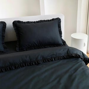Könnte beinhalten: Schwarzes Bettwäsche-Set mit einem Kissen und einem Bettbezug mit Rüschenkanten. Das Kissen ist rechteckig und mittig auf dem Bett platziert. Der Bettbezug ist ordentlich angeordnet, wobei die Rüschendetails am Rand sichtbar sind. Das Set besteht aus einem glatten, schwarzen Stoff.