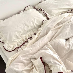 May include: A cream-coloured bedding set featuring two rectangular pillowcases and a duvet cover, all adorned with ruffled edges trimmed in a deep burgundy red piping. The fabric appears smooth and lightweight, covering a bed with a white headboard.