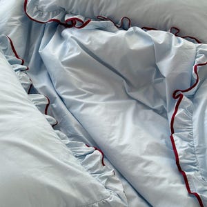 May include: Light blue bedding set with ruffled edges. The set includes a pillow and a duvet cover, both trimmed with a dark red border. The fabric appears soft and slightly wrinkled, suggesting a comfortable texture.