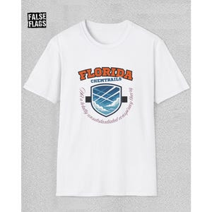 Puede incluir: Camiseta blanca con un diseño que dice "FLORIDA CHEMTRAILS" en naranja y azul. El diseño incluye un escudo con nubes y cielo, y el texto "Not a totally unsubstantiated conspiracy theory".