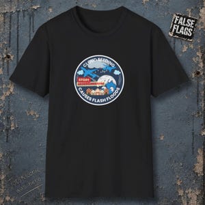 May include: A black t-shirt featuring a circular graphic with two blue airplanes, a large wave, and houses. The text reads: CLOUD SEEDING, STOP! GEOENGINEERING, CAUSES FLASH FLOODS. A small rectangular patch in the top right corner displays the text FALSE FLAGS.