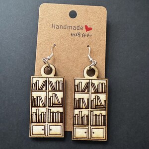 May include: A pair of wooden earrings shaped like bookshelves with books on the shelves. The earrings are brown and have silver hooks. The tag says "Handmade with love".