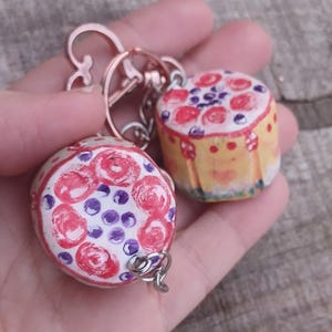 May include: Two handmade keyrings with miniature cake designs. One keyring has a white base with red roses and purple dots. The other keyring has a yellow base with red roses and purple berries. Both keyrings have a metal ring.